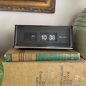May include: A black Bulova flip clock displaying the time as 10:38. The clock sits atop two vintage books with gold lettering and a green book. The clock has a silver trim and the word "Bulova" is printed on the front.