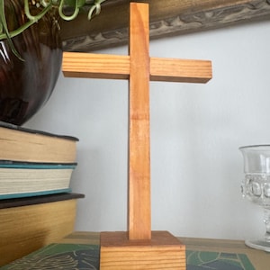 May include: A wooden cross with a natural finish, standing upright on a small square base. The cross is made of light-colored wood and has a simple, traditional design. The cross is approximately 10 inches tall.