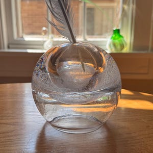 May include: A clear, spherical glass vase with a feather. The vase has a textured, bubble-like design and sits on a wooden surface. A small green glass vase is visible in the background, near a window.