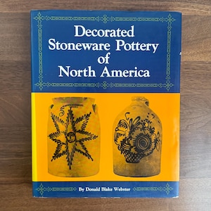 May include: A book titled "Decorated Stoneware Pottery of North America" with a blue and yellow cover. The cover features two antique stoneware pottery pieces with black designs. The author's name, Donald Blake Webster, is also visible.