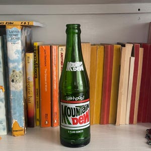 May include: A green Mountain Dew glass bottle with a white and red label, featuring the text "Yahoo!" and "Mountain Dew". The bottle contains 8 fluid ounces. The bottle is on a shelf with a collection of books.