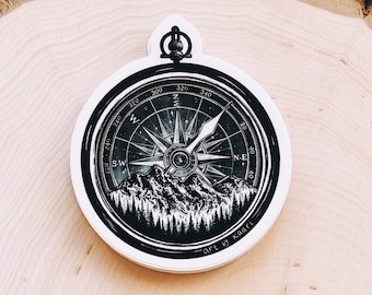 May include: A black and white compass sticker with a mountain and forest design. The compass rose features directional markings and a needle. The sticker has the text "art by kaari".
