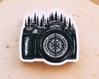 May include: Black and white illustration of a camera with a compass in the lens. The camera is surrounded by a forest of trees.