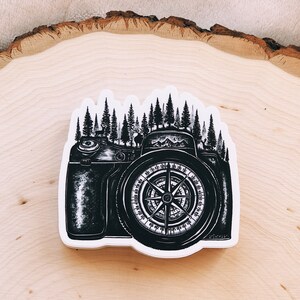 May include: Black and white illustration of a camera with a compass in the lens. The camera is surrounded by a forest of trees.