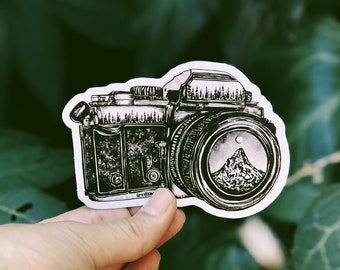 May include: A black and white illustration of a vintage camera with a mountain scene in the viewfinder. The camera has a textured surface and a lens with a silver rim.