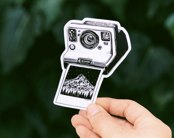 May include: Black and white sticker of a vintage instant camera with a mountain landscape photo. The camera is detailed with a lens, flash, and buttons. The photo shows a mountain range, trees, and a starry sky.