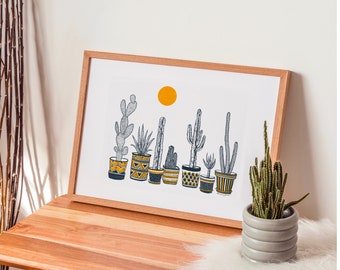 Op de afbeelding: Een ingelijste kunstprint met een rij cactusplanten in potten met een gele zon tegen een witte achtergrond. De potten hebben geometrische patronen in geel, zwart en wit. Een kleine cactus in een grijze pot staat ernaast op een houten tafel.