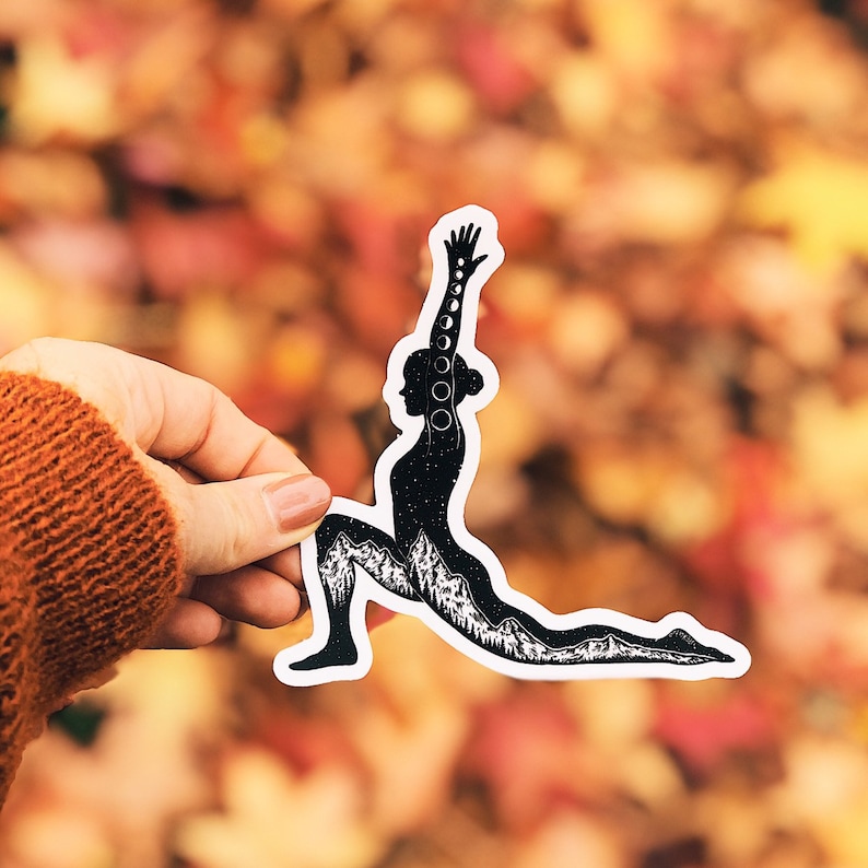 May include: Black and white silhouette sticker of a person in a yoga pose, with a mountain range and moon phases incorporated into the design. The sticker is on a background of fall leaves.