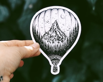 May include: Black and white sticker featuring a hot air balloon design. The balloon's interior showcases a mountain range, forest, moon, and birds. The sticker is held against a blurred green background.