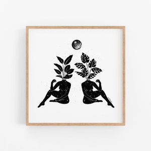May include: Black and white art print featuring two figures with plant-like heads, seated in a relaxed pose. A full moon is positioned above them. The artwork is framed in a light-colored wooden frame.