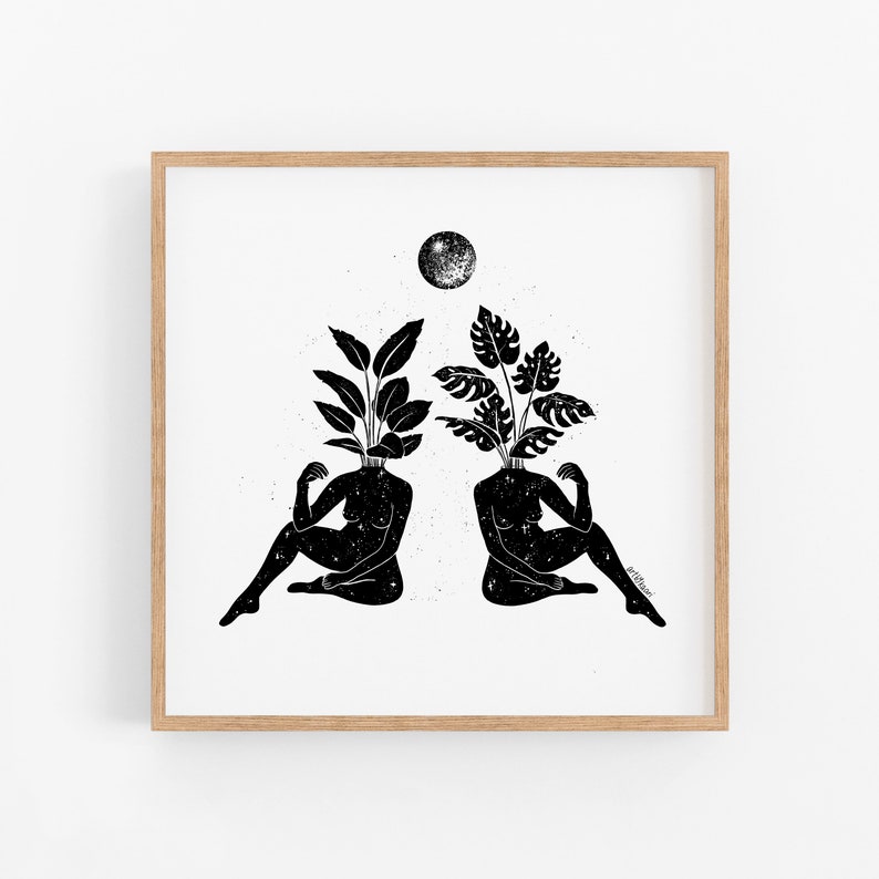 May include: Black and white art print featuring two figures with plant-like heads, seated in a relaxed pose. A full moon is positioned above them. The artwork is framed in a light-colored wooden frame.