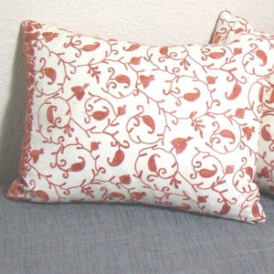 May include: Two white throw pillows with a red floral embroidery pattern. The pillows are on a gray couch.
