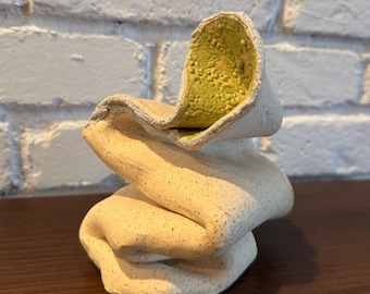 Puede incluir: Una escultura de cerámica artesanal con un diseño abstracto único. El exterior es de color blanco roto moteado, mientras que el interior tiene un acabado texturizado amarillo-verdoso. La forma es orgánica y fluida, que se asemeja a tela doblada. La escultura se exhibe sobre una superficie marrón oscuro.