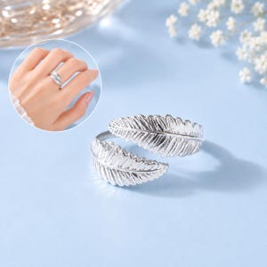 May include: Two silver feather rings. One ring is on a person's finger, and the other is displayed separately. The rings have a detailed feather design and are set against a light blue background.