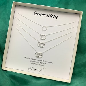 Puede incluir: Una caja blanca con un collar de plata con cuatro círculos entrelazados. La caja tiene el texto "Generations" y "The love between a Great-Grandmother, Grandmother, Mother & daughter is forever" impreso en ella.
