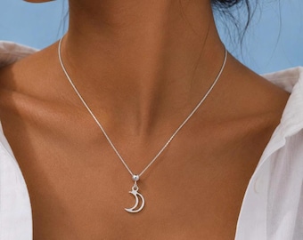May include: A silver-toned necklace featuring a delicate chain and a small, polished crescent moon pendant. The pendant hangs from a simple circular bail, creating a clean and understated look that rests against the collarbone area.