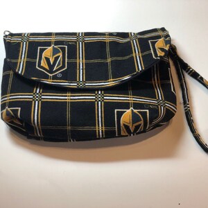 May include: A black and gold fabric clutch purse with a plaid pattern and the NHL Vegas Golden Knights logo. The purse has a short strap.