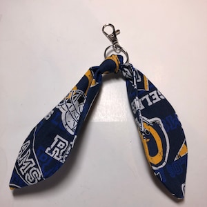 May include: A blue and yellow fabric keychain with a silver clip. The fabric has a repeated pattern of the Los Angeles Rams logo and the words "Rams" and "Los Angeles".