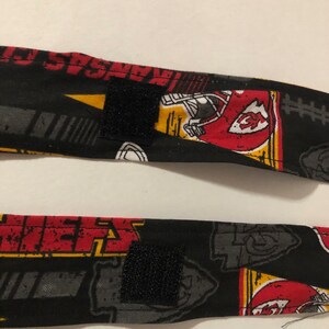 May include: Two black fabric straps with a red, yellow, and white pattern featuring the Kansas City Chiefs logo and the words "Kansas City Chiefs". The straps have black hook and loop fasteners.