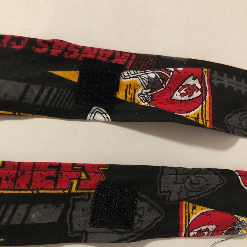 May include: Two black fabric straps with a red, yellow, and white pattern featuring the Kansas City Chiefs logo and the words "Kansas City Chiefs". The straps have black hook and loop fasteners.