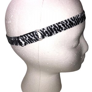 May include: A black and white zebra print headband is displayed on a white foam head. The headband is made of a thin, flexible material and features a repeating pattern of black stripes on a white background.
