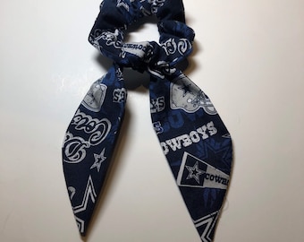 May include: A blue and white hair scrunchie with a Dallas Cowboys theme. The scrunchie has a long tail with the Cowboys logo and the words "Cowboys" and "Dallas" on it.