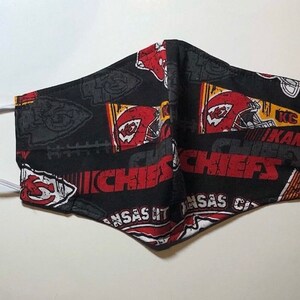 May include: A black face mask with a white elastic band. The mask is printed with a red, yellow, and white pattern of the Kansas City Chiefs logo and the words "Kansas City Chiefs".