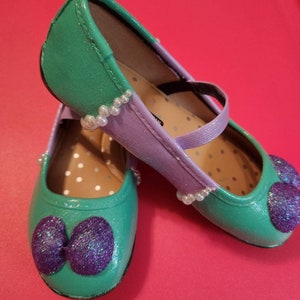 May include: A pair of green and purple glitter shoes with a pearl trim. The shoes have a bow on the toe and a strap across the top.