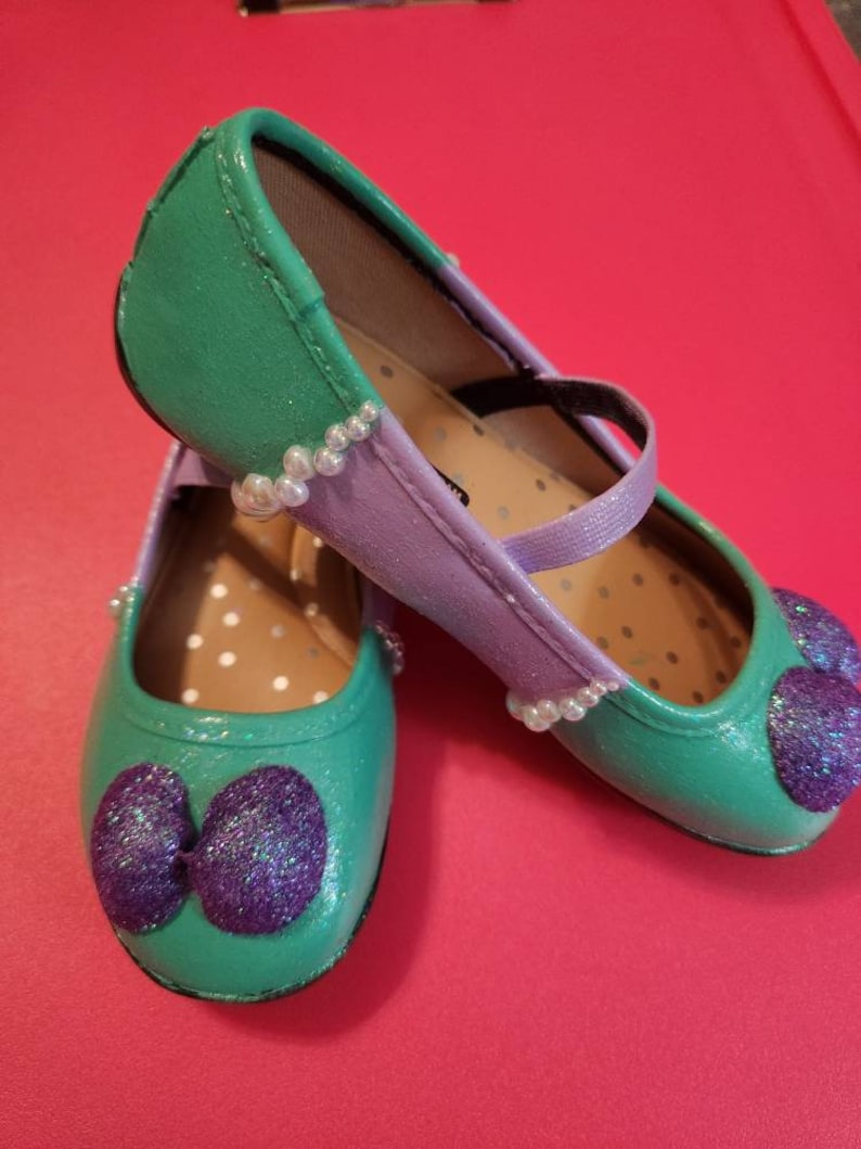 May include: A pair of green and purple glitter shoes with a pearl trim. The shoes have a bow on the toe and a strap across the top.
