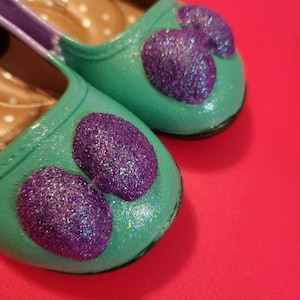 May include: A pair of teal blue children's shoes with purple glitter hearts. The shoes have a purple strap and a white polka dot lining.
