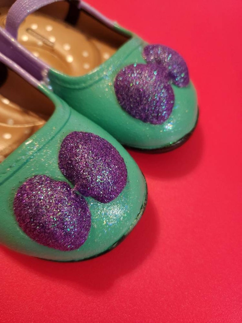 May include: A pair of teal blue children's shoes with purple glitter hearts. The shoes have a purple strap and a white polka dot lining.