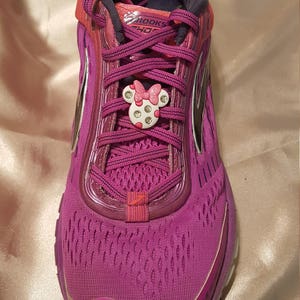 May include: Purple running shoes with pink laces and a pink Minnie Mouse shoe charm. The shoes have a black and white Brooks logo on the side.