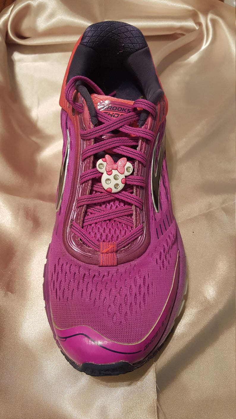 May include: Purple running shoes with pink laces and a pink Minnie Mouse shoe charm. The shoes have a black and white Brooks logo on the side.