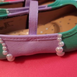 May include: A pair of purple and green children's shoes with a pearl trim. The shoes have a strap across the top and a pointed toe.