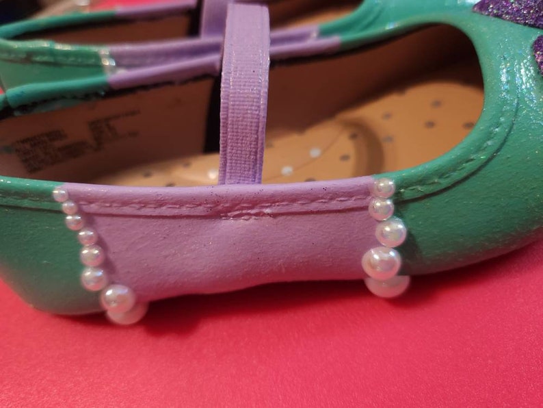 May include: A pair of purple and green children's shoes with a pearl trim. The shoes have a strap across the top and a pointed toe.
