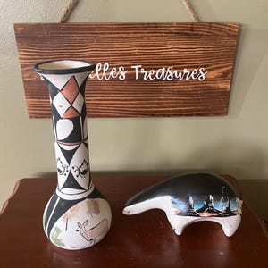 May include: A tall, decorative vase with a black, white, and orange geometric design, and a small ceramic bear figurine with a landscape design. The vase has a deer illustration. A wooden sign with the text "elles Treasures" hangs in the background.