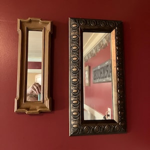 May include: Two rectangular mirrors with ornate frames hang on a red wall. The mirror on the left has a light brown frame with a simple design. The mirror on the right has a dark brown frame with an oval pattern.
