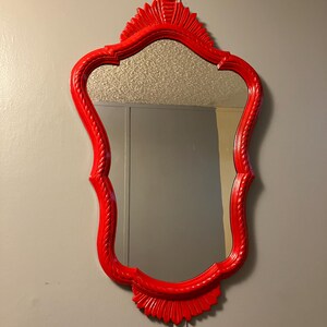 May include: A wall-mounted mirror featuring a vibrant, glossy red frame with an ornate, scalloped edge and decorative carved detailing at the top and bottom. The reflective glass surface is clear and smooth, framed by a raised, rope-like texture.