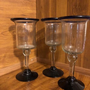 May include: Three clear glass candle holders with black bases and a blue rim. The candle holders are on a wooden surface.