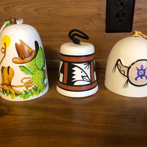 May include: Three ceramic bells with Southwestern designs. One bell features a cowboy hat, cactus, and boots. Another has a geometric pattern with an eagle. The third bell displays a turtle symbol. Each bell has a rope or cord for hanging.