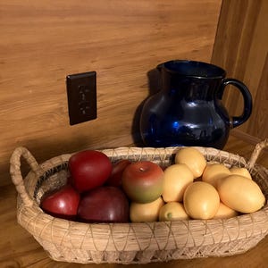May include: A still life arrangement featuring a woven basket filled with red and yellow apples and lemons. A dark blue pitcher sits behind the basket. The scene is set on a wooden surface.