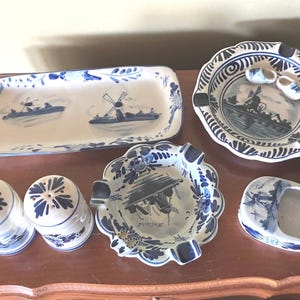 May include: A set of blue and white Dutch Delftware pottery, including a rectangular serving tray with a windmill design, two small salt and pepper shakers, and three ashtrays with floral designs. The ashtrays feature a blue and white floral pattern with a central image of a windmill.