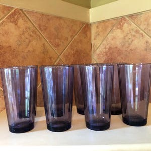 May include: Six purple glass tumblers with a textured surface. The glasses are arranged in a row on a tiled countertop.