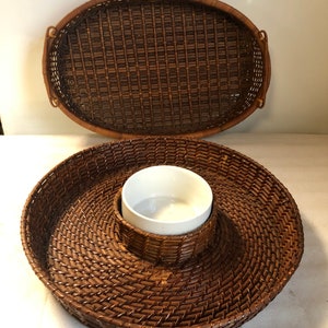 May include: A set of two brown wicker serving trays. The larger tray is oval shaped and the smaller tray is round with a white ceramic bowl in the center.