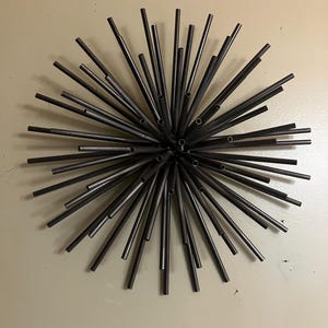 May include: A dark gray metal starburst wall sculpture featuring numerous thin, hollow cylindrical rods radiating outward from a central point, creating a spherical, spiky, and symmetrical sunburst design with a smooth, matte finish.