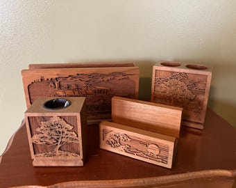 May include: A collection of wooden desk organizers with carved designs. Includes a pen holder, business card holder, and a letter holder. The designs feature a tree, a train, and a sunset scene. The wood has a warm, reddish-brown tone.