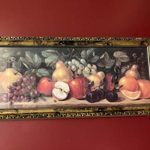 May include: A framed still life painting featuring a variety of fruits. The artwork includes red apples, grapes, pears, and oranges. The frame is gold-colored with ornate detailing, adding a classic touch to the piece.