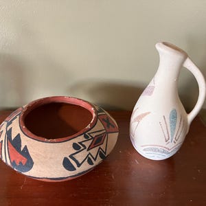 May include: Two handcrafted pottery pieces. A round bowl with geometric designs in red, black, and tan. Next to it, a small pitcher with a handle, decorated with pastel floral motifs. Both are on a dark wooden surface.