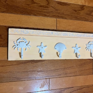 May include: A decorative wall-mounted coat rack with five hooks. The hooks are shaped like a crab, starfish, and seashell. The rack is painted light yellow with a decorative border and is mounted on a wooden surface.