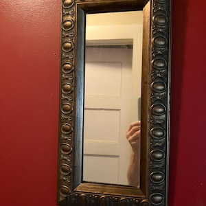 May include: Rectangular wall mirror with a decorative, ornate bronze-colored frame. The frame features a repeating oval and leaf design. The mirror is hanging on a red wall.